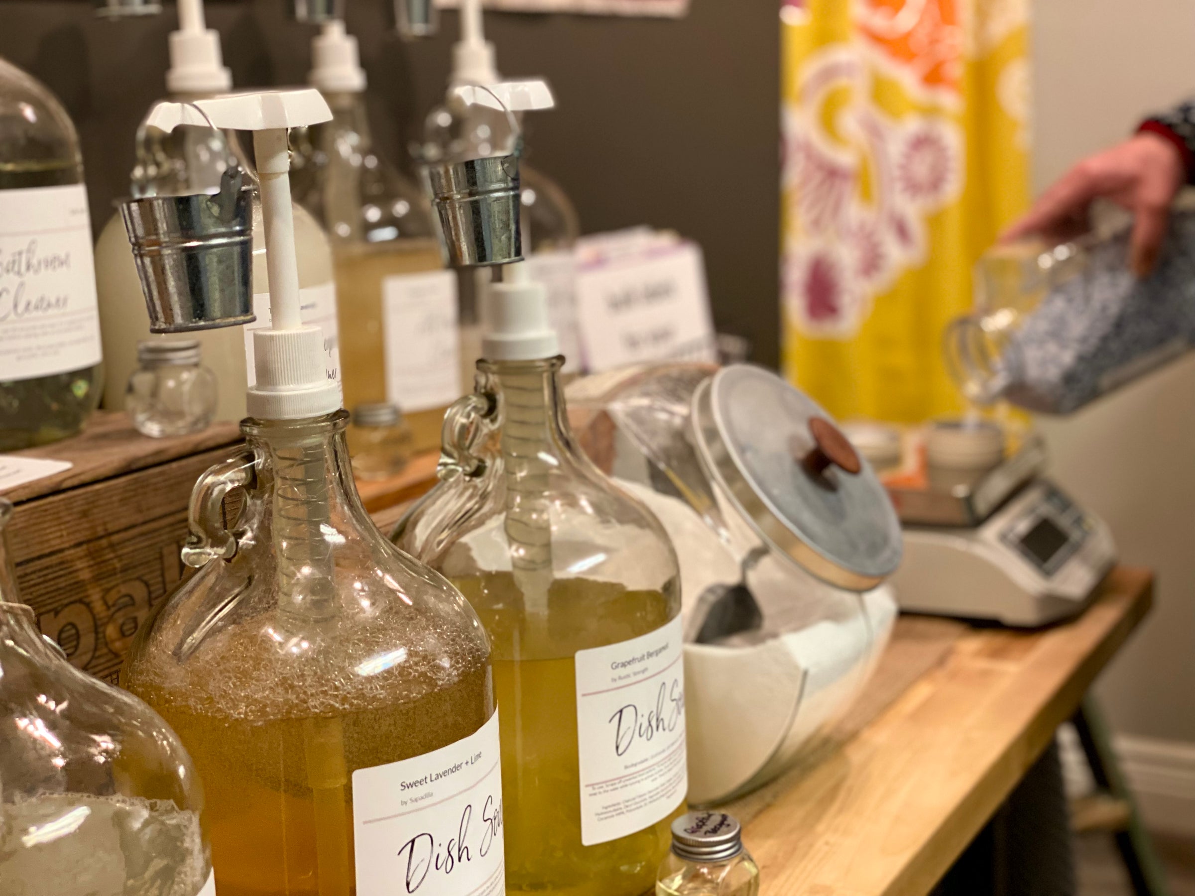 Glass dispensers labeled Dish Soap sit on a wooden counter near jars of powder, a scale, and a person pouring powder from a jar in the background. The setting appears to be a refill or zero-waste store.