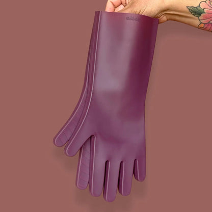 A hand with a floral tattoo holds up a pair of long, purple Keeps Silicone Cleaning Gloves against a matching solid background.