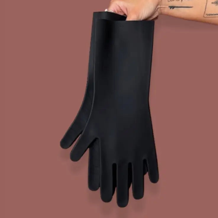 A hand with tattoos holds a pair of Keeps Silicone Cleaning Gloves against a solid brown background.