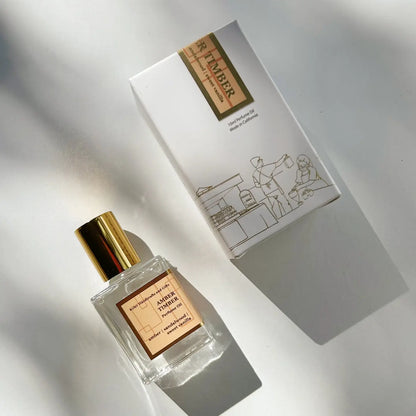 A bottle of Kyler Handcrafts Roll On Perfume, featuring a phthalate-free formula and topped with a gold cap, rests beside its box adorned with a minimalist line drawing of people and objects. Sunlight streams in, casting gentle shadows and highlighting the bright, elegant atmosphere.