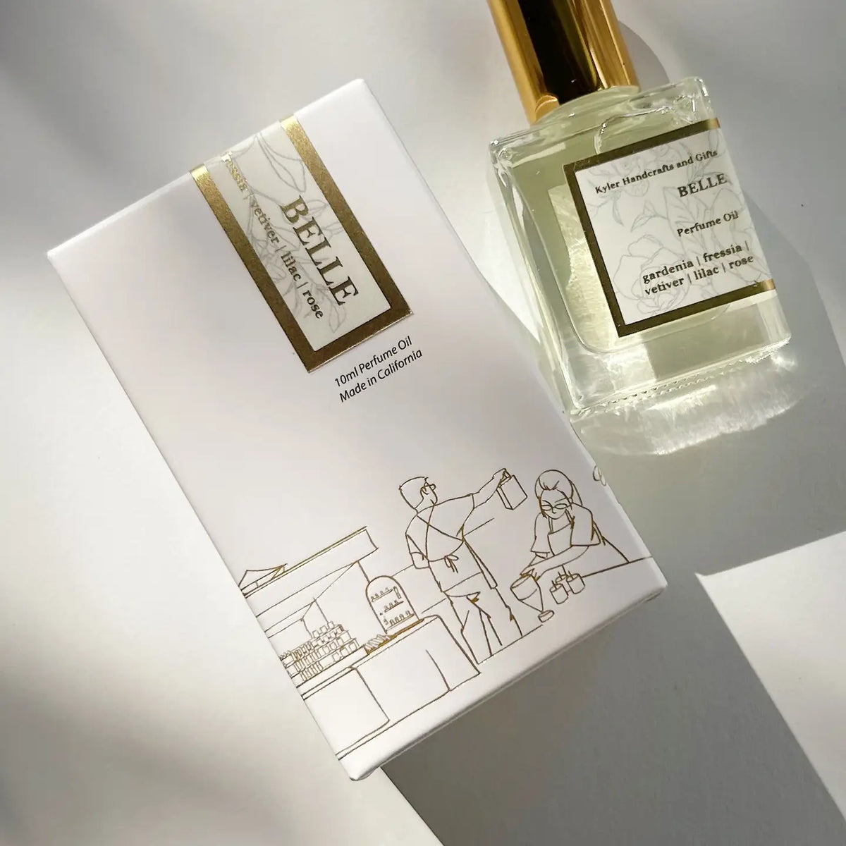 A bottle of Kyler Handcrafts Roll On Perfume with a gold cap rests beside its white box, adorned with a simple line drawing of two people at a market stand. Sunlight casts soft shadows across both items, highlighting this handcrafted perfume’s elegant and phthalate-free fragrance.