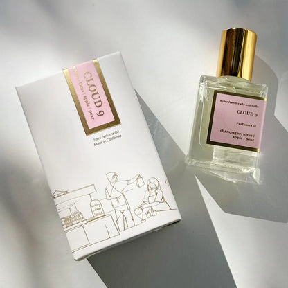 A bottle of Kyler Handcrafts Roll On Perfume with a gold cap rests next to its white and pink box, adorned with gold accents and an illustration of people at a café. This phthalate-free fragrance delivers the luxurious experience of roll-on perfume oils in a stylish presentation.