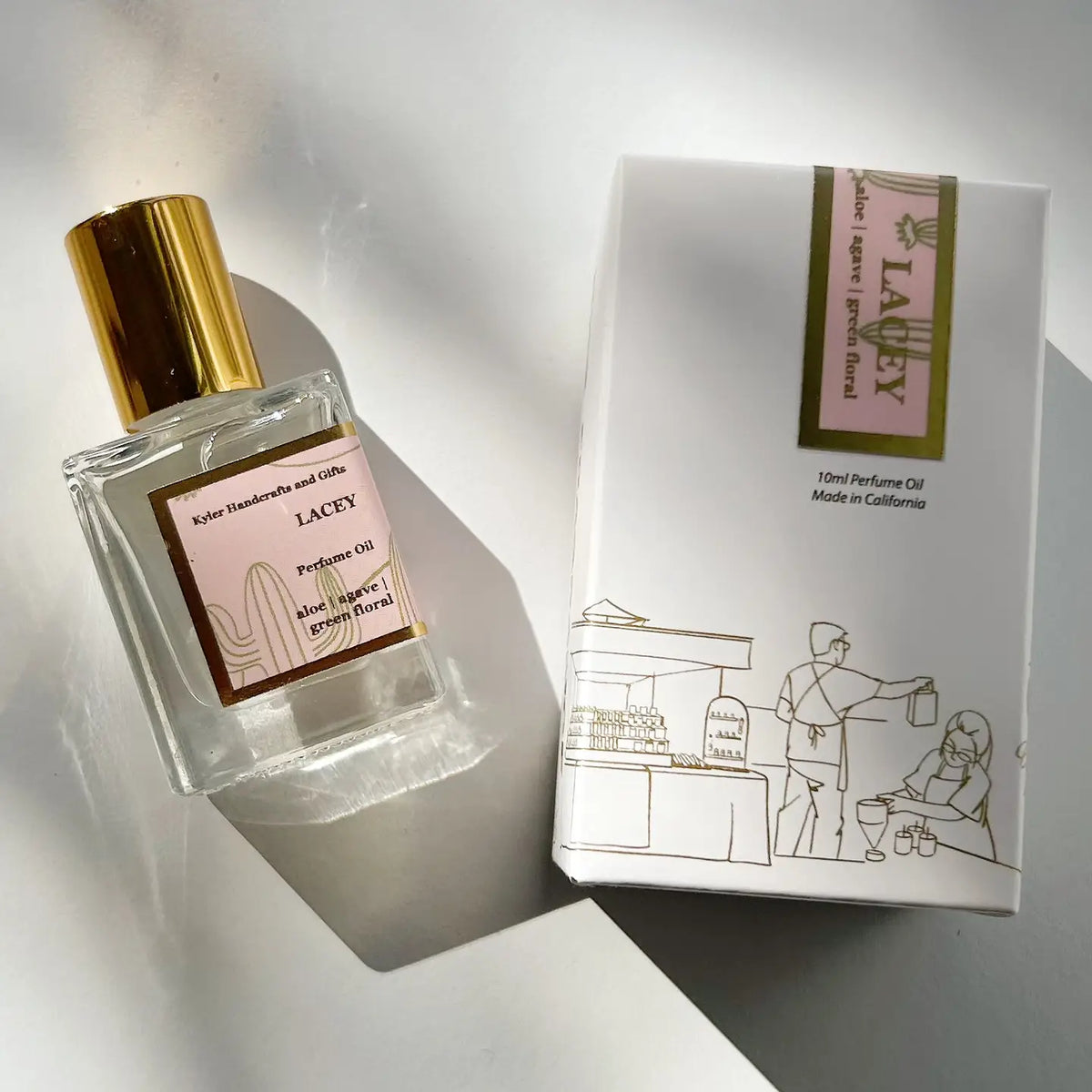 A glass roll on perfume bottle with a gold cap, labeled “Roll On Perfume” by Kyler Handcrafts, sits beside its white box adorned with a gold and pink label and an illustration of a person handing a drink to a woman at a booth. This handcrafted perfume is made with phthalate-free fragrance, providing an elegant experience for those who value high-quality scents.