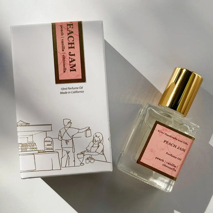 A bottle of Kyler Handcrafts Roll On Perfume in the Peach Jam scent, topped with a gold cap, is positioned beside its box adorned with a minimal line drawing of a café scene. Both the bottle and box are labeled “Peach Jam: peach, vanilla, citronella.” This handcrafted roll-on perfume from Kyler Handcrafts is made using phthalate-free fragrance for a pure and delightful scent experience.