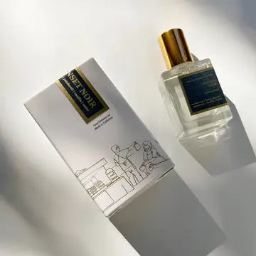 A handcrafted Roll On Perfume by Kyler Handcrafts, featuring a gold-capped bottle, rests next to its box adorned with minimalist line art and a dark blue and gold label. Both items cast soft shadows on a white surface, highlighting the elegance of this phthalate-free fragrance.