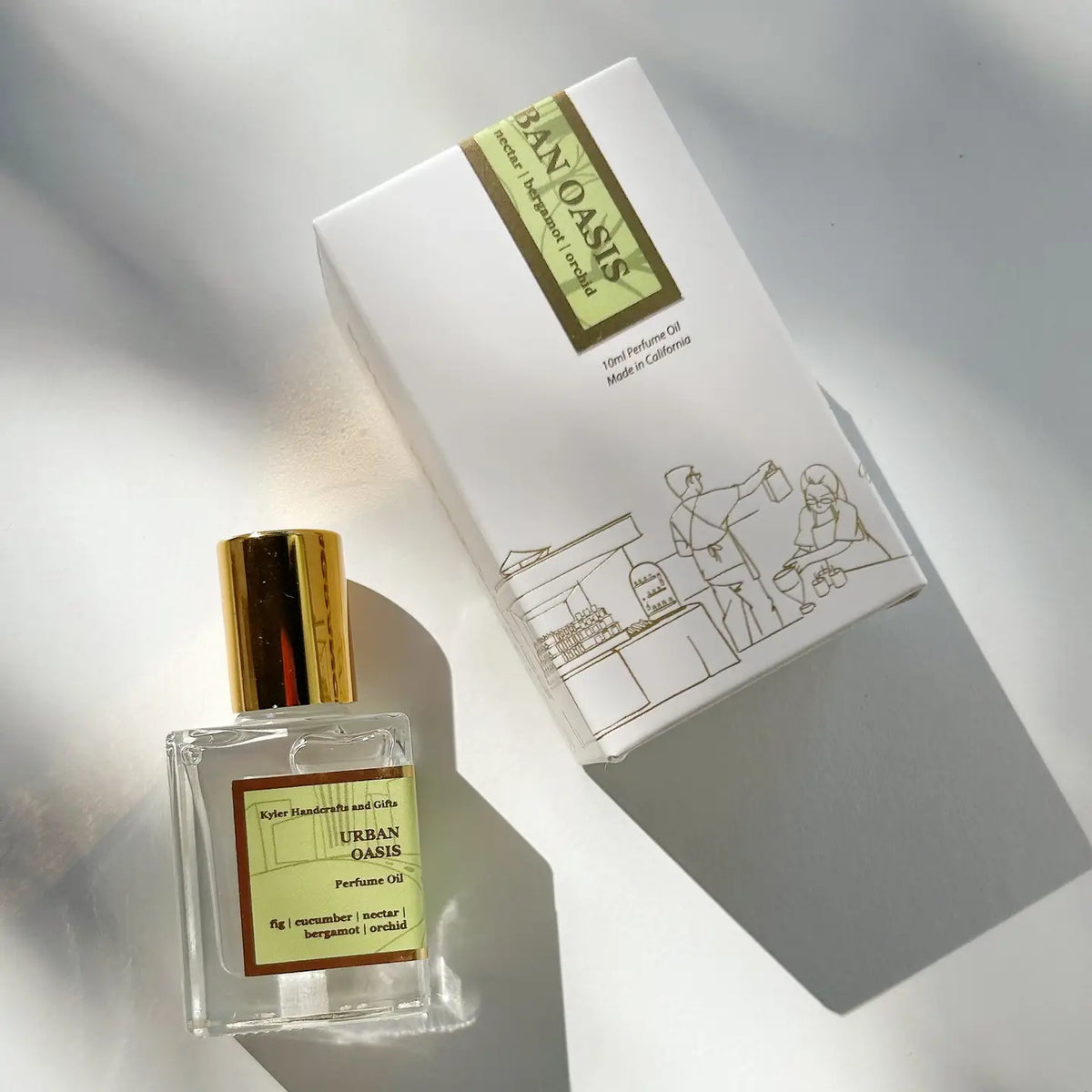 A bottle of Kyler Handcrafts Roll On Perfume with a gold cap rests beside its white box, adorned with an illustrated street vendor scene. This handcrafted, phthalate-free fragrance is bathed in soft sunlight, casting gentle shadows across the white surface.