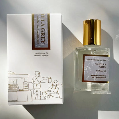 A clear Roll On Perfume bottle with a gold cap, labeled Vanilla Grey by Kyler Handcrafts, sits beside its white box adorned with an illustration of two people at a market table. Sunlight casts gentle shadows across both items, highlighting the allure of this phthalate-free fragrance—carefully crafted by Kyler Handcrafts for a pure and elegant scent experience.