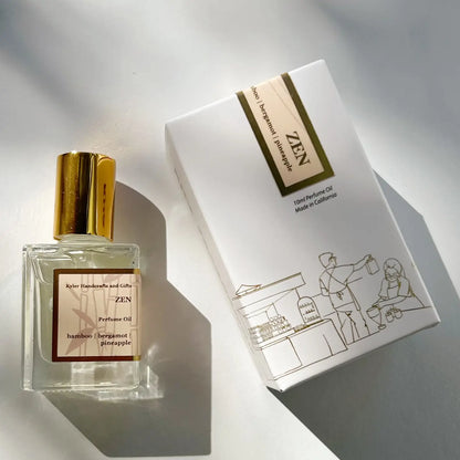 A glass perfume bottle with a gold cap labeled “Roll On Perfume” by Kyler Handcrafts sits beside a white box decorated with minimalist line art of people shopping. Sunlight and shadows play across both items, accentuating the elegance of this handcrafted perfume made with phthalate-free fragrance.