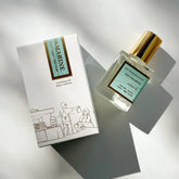 A Kyler Handcrafts Roll On Perfume with a gold cap rests by its illustrated white box with gold details, bathed in soft natural light. Phthalate-free, this handcrafted fragrance exudes elegance and charm.