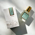 A Kyler Handcrafts Roll On Perfume with a gold cap rests by its illustrated white box with gold details, bathed in soft natural light. Phthalate-free, this handcrafted fragrance exudes elegance and charm.