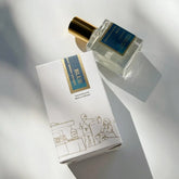 A bottle of Kyler Handcrafts Roll On Perfume with a gold cap rests next to its white box, which displays a blue and gold label and a minimalist market scene line drawing, all set on a bright surface with soft shadows.