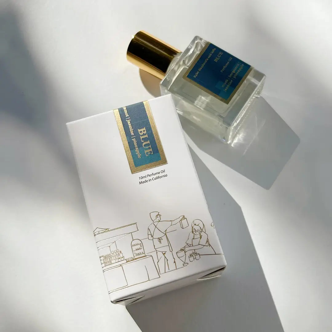 A bottle of Kyler Handcrafts Roll On Perfume with a gold cap rests next to its white box, which displays a blue and gold label and a minimalist market scene line drawing, all set on a bright surface with soft shadows.