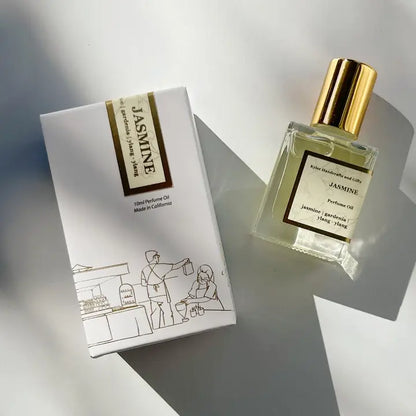 A bottle of Kyler Handcrafts Roll On Perfume with a gold cap sits beside its white box, featuring a minimalist line drawing of two people at a market stall. Sunlight creates gentle shadows on the light surface.