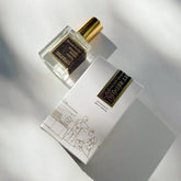 A handcrafted Kyler Handcrafts Roll On Perfume with a gold cap rests by a white box adorned with minimalist line art and a dark, gold-accented label. Both cast soft shadows—a phthalate-free fragrance experience.