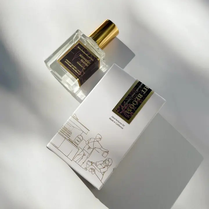 A handcrafted Kyler Handcrafts Roll On Perfume with a gold cap rests by a white box adorned with minimalist line art and a dark, gold-accented label. Both cast soft shadows—a phthalate-free fragrance experience.