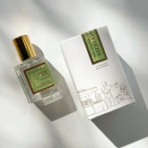 A glass bottle of Kyler Handcrafts Roll On Perfume with a gold cap labeled Sunday Olive rests beside a white box adorned with minimalist line art of people shopping outdoors, both softly illuminated by natural sunlight.
