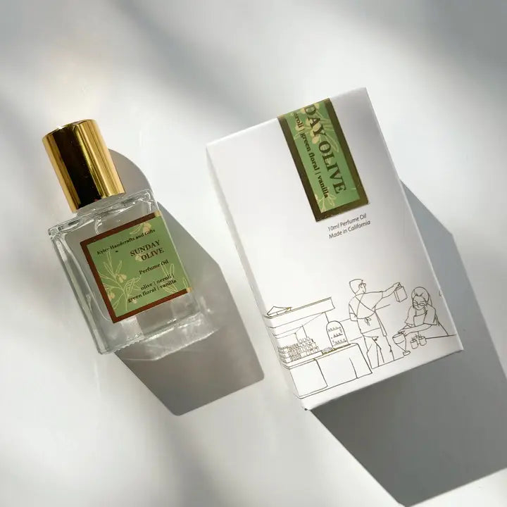 A glass bottle of Kyler Handcrafts Roll On Perfume with a gold cap labeled Sunday Olive rests beside a white box adorned with minimalist line art of people shopping outdoors, both softly illuminated by natural sunlight.
