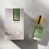 A bottle of Kyler Handcrafts Roll On Perfume with a gold cap sits beside its white box, decorated with a minimalist line drawing of two people at a market stall. Sunlight casts gentle shadows on this handcrafted scent.