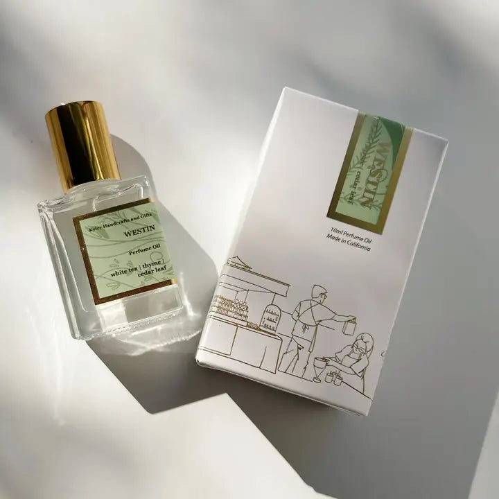 A bottle of Kyler Handcrafts Roll On Perfume with a gold cap sits beside its white box, adorned with minimalist line art of two people and green-gold accents, both softly illuminated by sunlight.