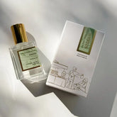 A bottle of Kyler Handcrafts Roll On Perfume with a gold cap sits beside its white box, adorned with minimalist line art of two people and green-gold accents, both softly illuminated by sunlight.
