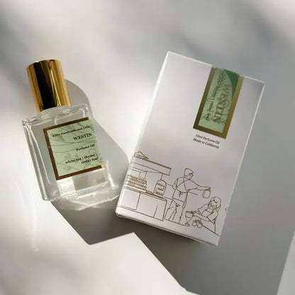 A bottle of Kyler Handcrafts Roll On Perfume with a gold cap sits beside its white box, adorned with minimalist line art of two people and green-gold accents, both softly illuminated by sunlight.