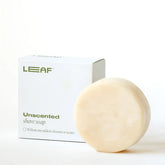 A round, pale Shave Soap Bar by Leaf sits next to a white box labeled "Leaf Shave Soap Bar Unscented," emphasizing its vegan formula with no added colorants or scents.