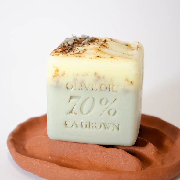 The Lianna Handmade Bar Soap by Lianna, embossed with "OLIVE OIL 70% CA GROWN," is a square, pale green and cream organic soap topped with dried herbs, displayed on a wavy, round reddish-brown dish.