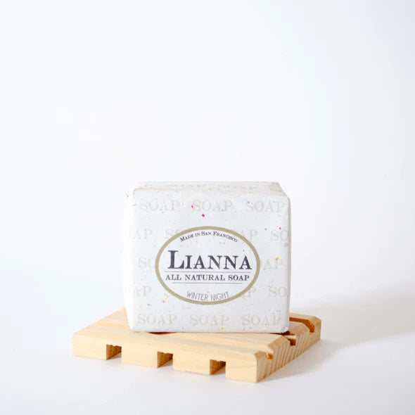 A white Lianna Handmade Bar Soap labeled "Winter Night" sits on a light wooden soap dish against a plain white background.