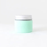 A mint green Protective Silicone Sleeve by Little Seed Farm rests on a plain white background. The sleeve, a sustainable alternative, features subtle embossed text on its side and fits snugly around jars.