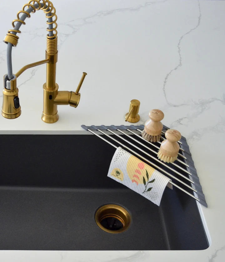 Kitchen sink with gold faucet, dish rack, and cleaning supplies on a marble countertop.