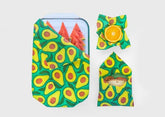 Three foods—a sandwich, watermelon slices in a container, and a halved orange—are wrapped in Meli Wraps Beeswax Wraps (3 pk) with an avocado design, highlighting stylish eco-friendly storage against a plain light background.