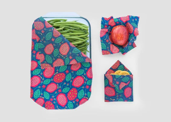 Meli Wraps Beeswax Wraps - 3 pk, featuring a blue and red fruit pattern, cover green beans, wrap an apple, and hold banana slices—an eco-friendly food storage solution shown against a light gray background.