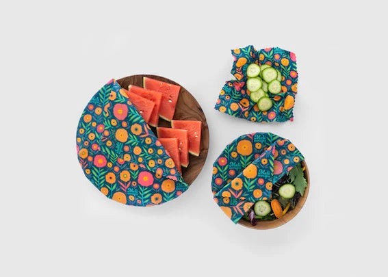 Three wooden bowls, each topped with Meli Wraps Beeswax Wraps - 3 pk in colorful floral patterns, keep watermelon slices, cucumber slices, and assorted greens fresh. An eco-friendly food storage solution shown against a plain white background.