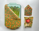 A glass container of asparagus covered with a green, eco-friendly Meli Wraps Beeswax Wrap sits beside two smaller Meli Wraps Beeswax Wraps—one folded, the other holding a red apple—on a white background.
