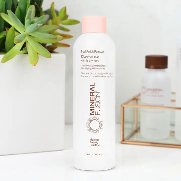 A white bottle of Mineral Fusion Acetone Free Nail Polish Remover sits on a white surface beside a green succulent. A blurred brown-capped bottle and gold wire rack appear in the background.
