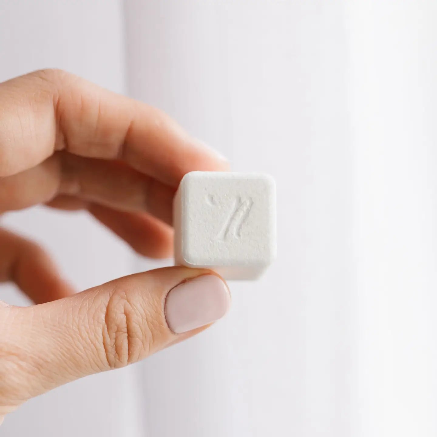 A hand with pale pink nail polish holds a small, white, square-shaped Laundry Cube from Nellie’s—with a subtle “7” embossed on its surface—against a blurred white background, highlighting the convenience and innovation of highly concentrated laundry detergent.