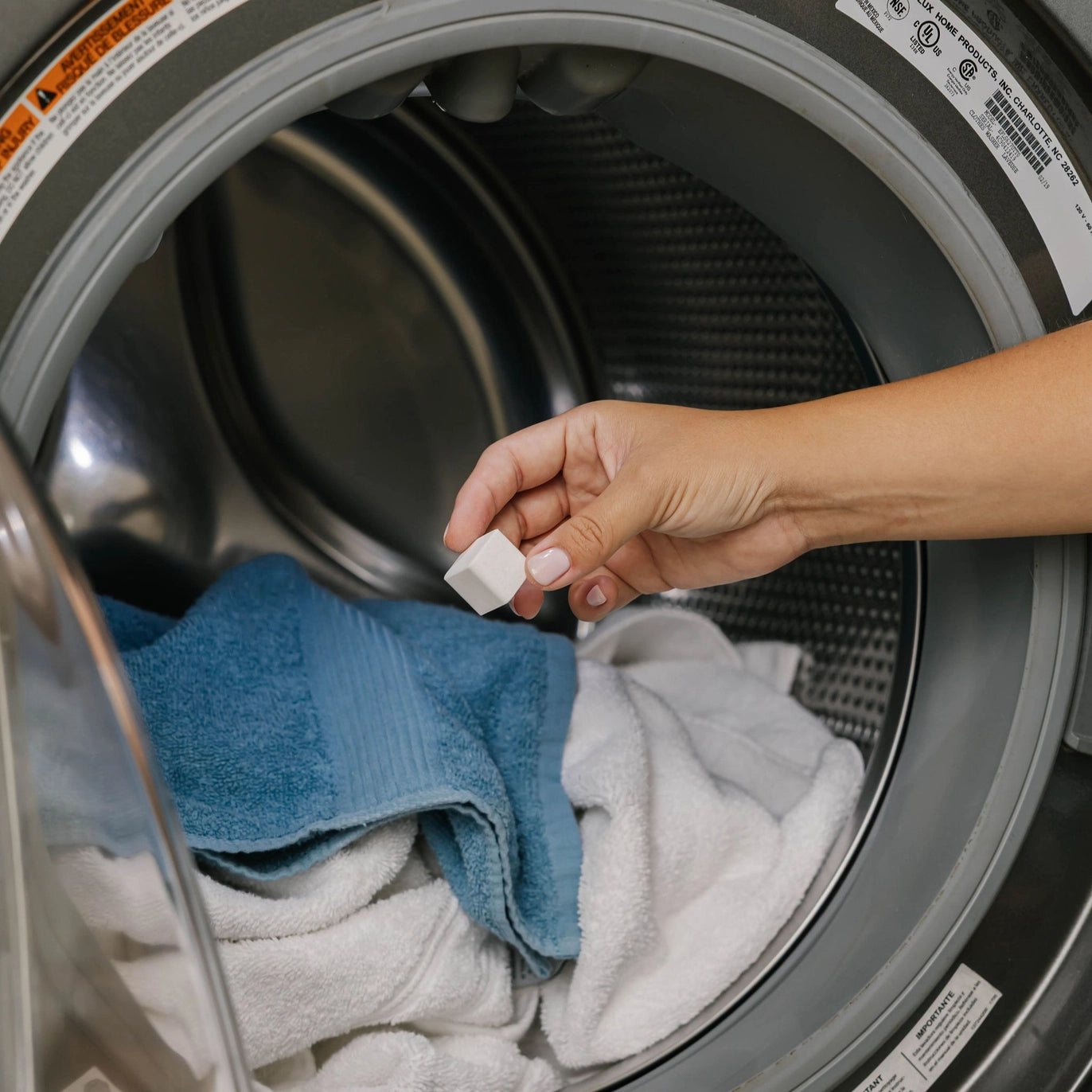 A hand holds a Nellie’s Laundry Cube, a highly concentrated detergent pod, above an open washing machine filled with towels, including a blue and a white one.