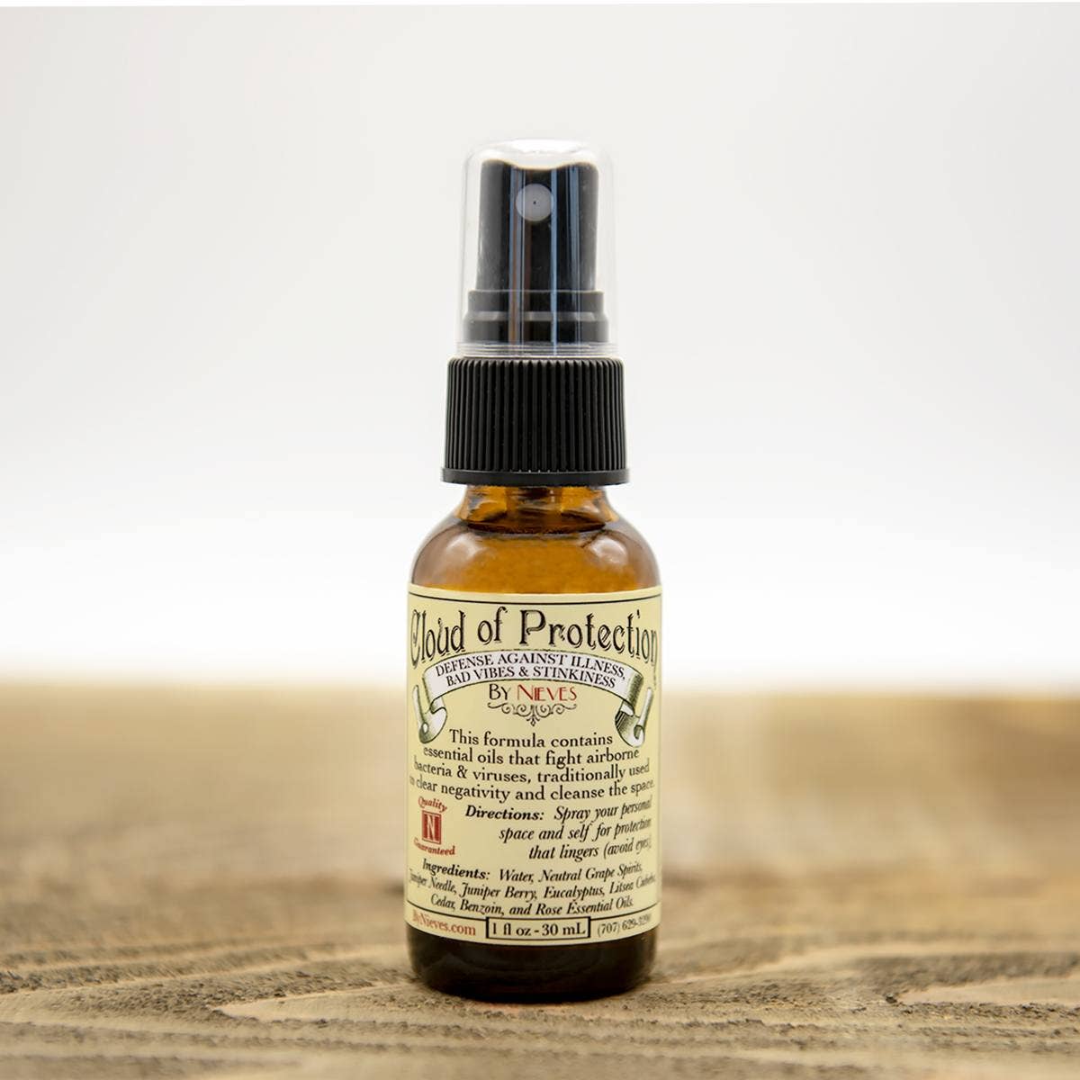 A small amber spray bottle labeled "Nieves Cloud of Protection" by Nieves rests on a wooden surface. The label highlights antimicrobial essential oils, making it a plant-based sanitizer with usage directions.