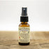 A small amber spray bottle labeled "Nieves Cloud of Protection" by Nieves rests on a wooden surface. The label highlights antimicrobial essential oils, making it a plant-based sanitizer with usage directions.