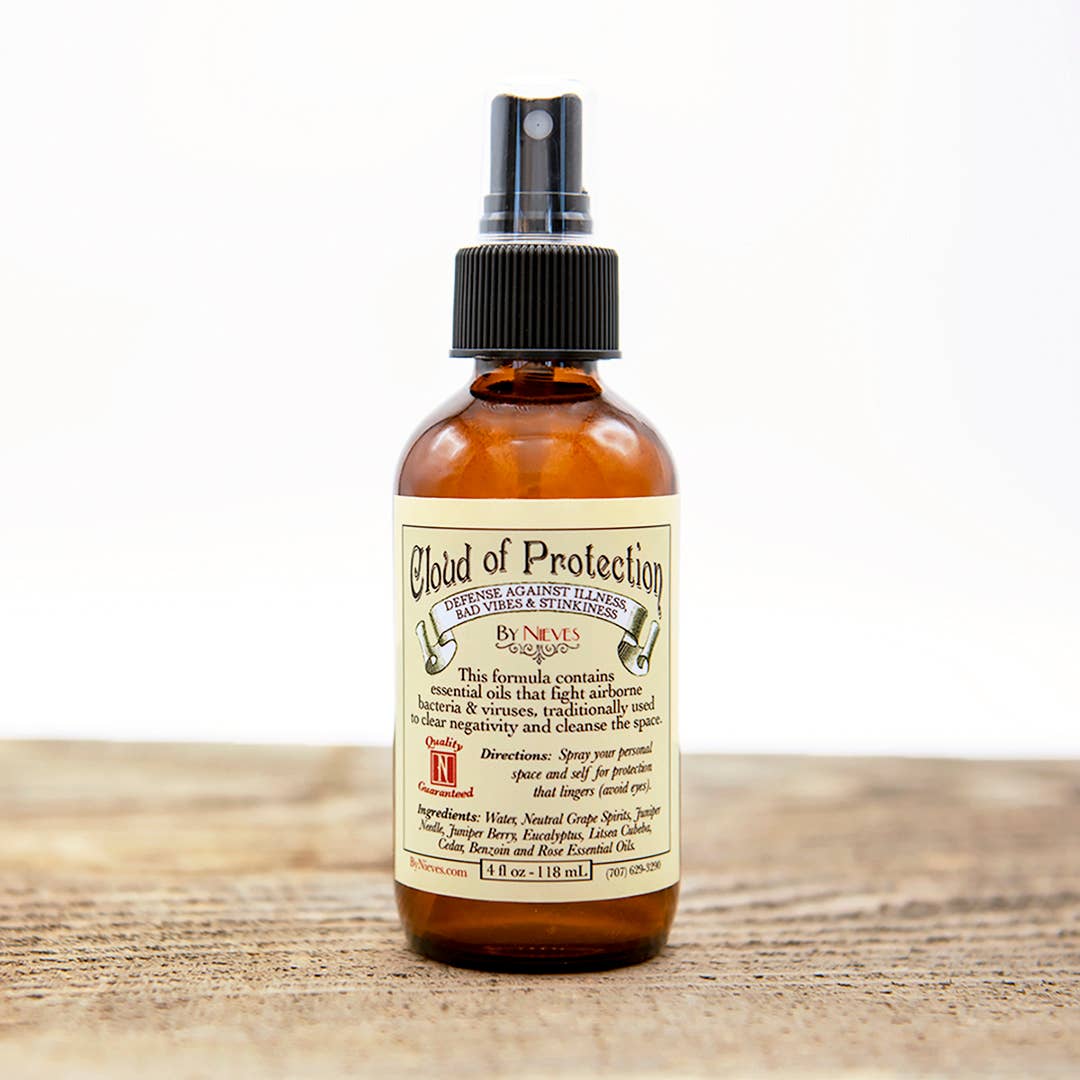 A brown glass bottle labeled "Nieves Cloud of Protection" sits on wood. The plant-powered sanitizer features antimicrobial essential oils and has a black spray nozzle. The Nieves brand product is set against a softly blurred white background.