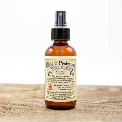 A brown glass bottle labeled "Nieves Cloud of Protection" sits on wood. The plant-powered sanitizer features antimicrobial essential oils and has a black spray nozzle. The Nieves brand product is set against a softly blurred white background.