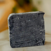 A close-up of the No Tox Life Underarm Detox Bar, a rectangular bar with textured grooves and lighter specks, stands upright on a light surface with a softly blurred background, emphasizing this natural skincare essential.