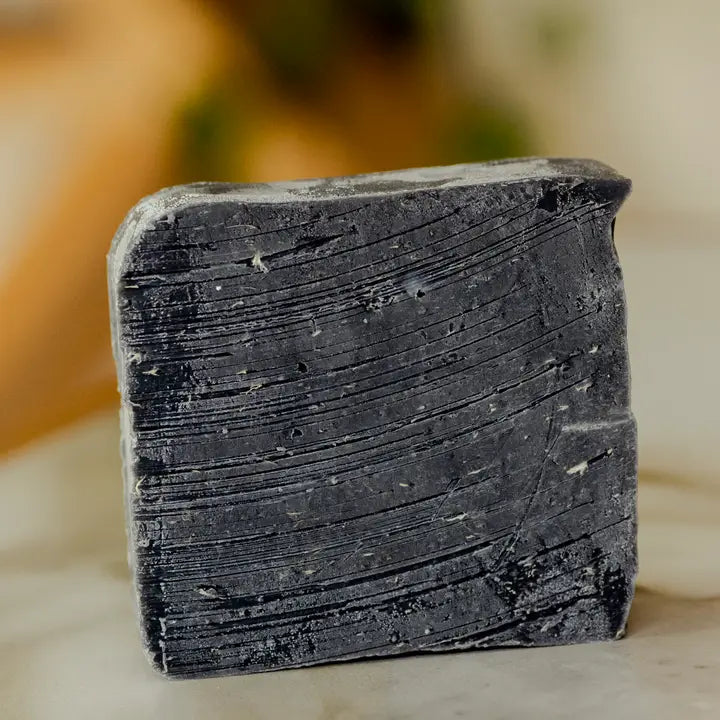 A close-up of the No Tox Life Underarm Detox Bar, a rectangular bar with textured grooves and lighter specks, stands upright on a light surface with a softly blurred background, emphasizing this natural skincare essential.