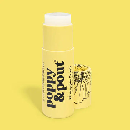 A yellow tube of Poppy &amp; Pout Lip Balm stands upright with the cap removed to reveal the balm inside. The cap, set next to it, displays a black floral illustration. The scene is set against a solid yellow background.