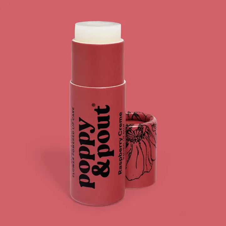 A tube of Poppy &amp; Pout Lip Balm stands upright with its cap off, showing the balm inside. The pink tube and cap feature black text, a floral illustration, and the Poppy &amp; Pout brand name.