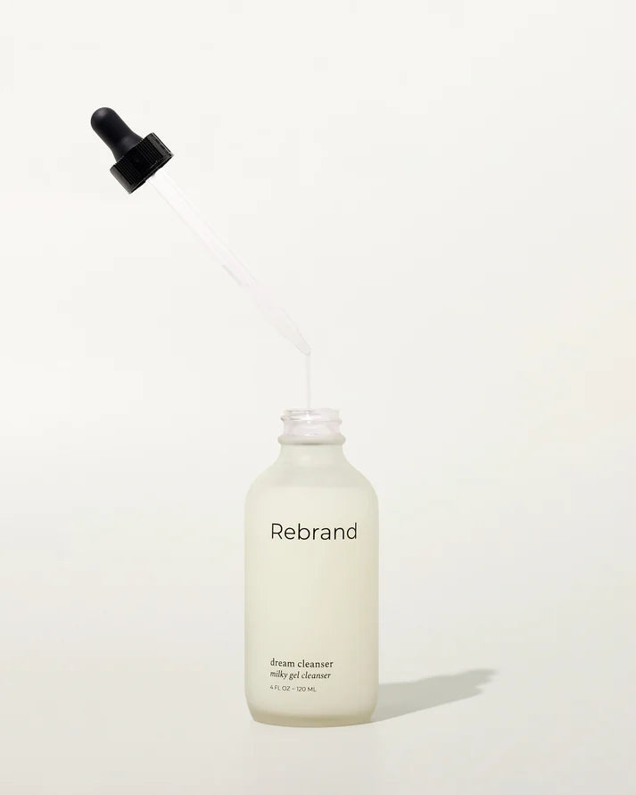 A clear glass bottle of Rebrand Dream Cleanser with a dropper rests on top, filled with light-colored liquid. The label highlights it as a milky gel cleanser and gentle makeup remover for sensitive skin. Background is plain and light.