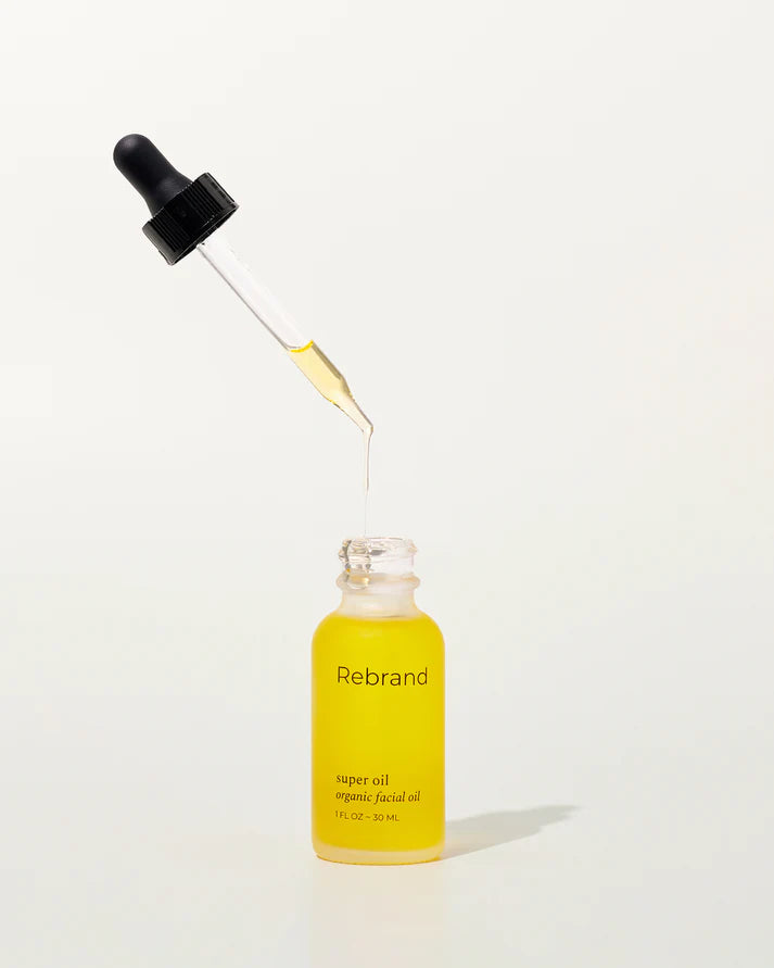 A dropper dispenses yellow oil above an open glass bottle labeled "Rebrand Super Oil"—a refillable facial oil crafted from organic plant oils—set against a plain light background.