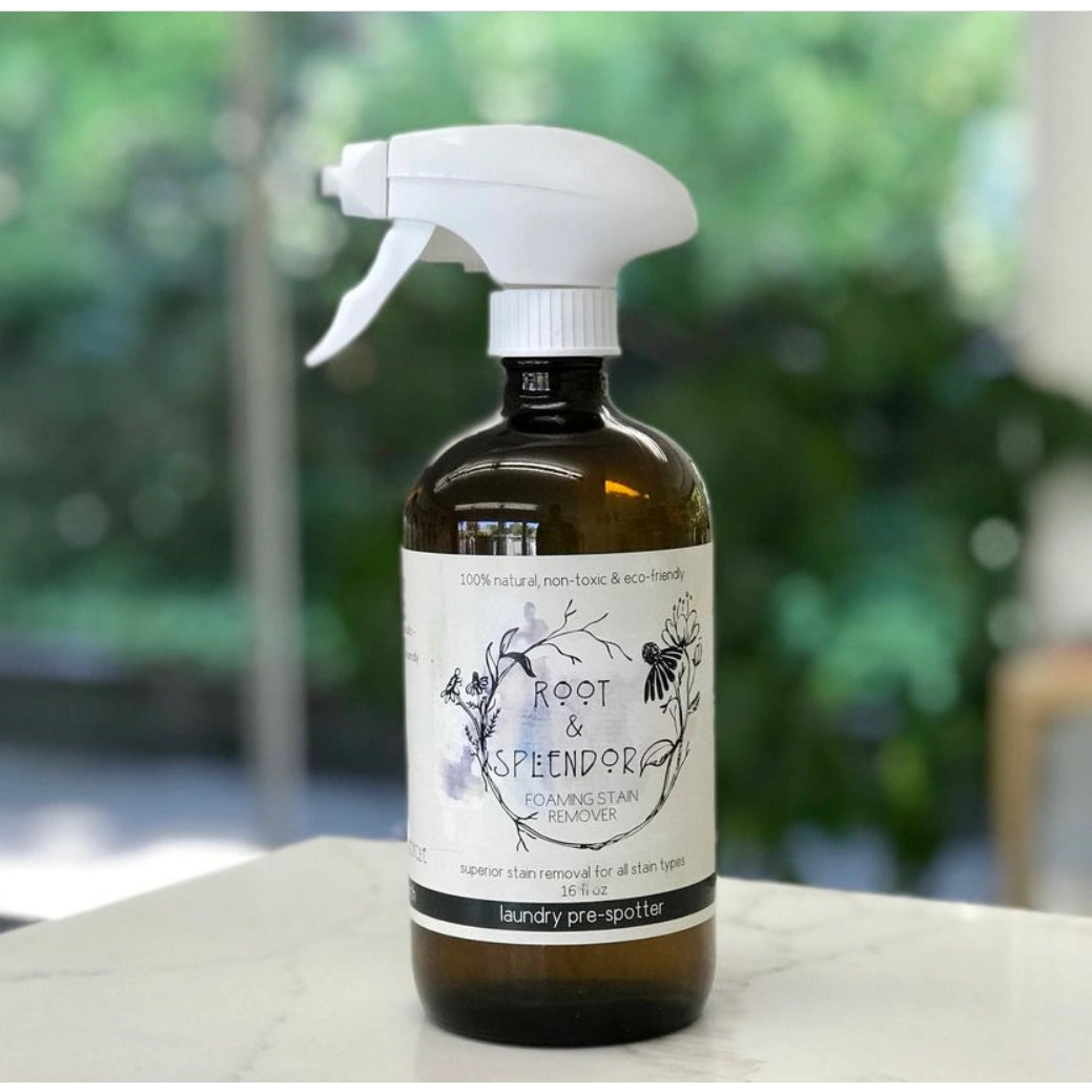 A brown spray bottle labeled Root and Splendor Laundry Pre-Spotter Stain Remover rests on a white surface with a blurred green background. This eco-friendly, natural, non-toxic laundry stain remover is safe for your home and the environment.