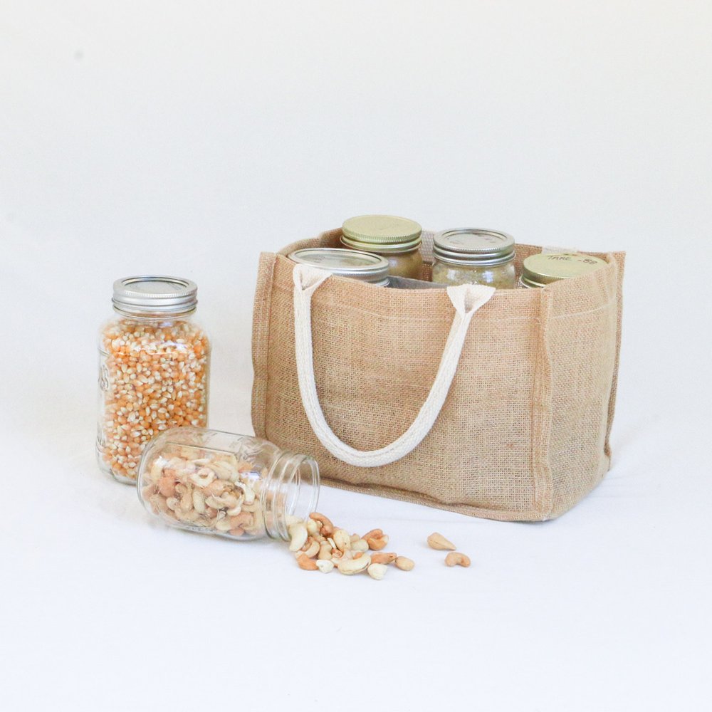 A Simple Ecology Reusable Jar Carrier with organic cotton handles sits on a white surface, holding jars with metal lids. One carrier contains popcorn kernels, while another lets cashews spill out.