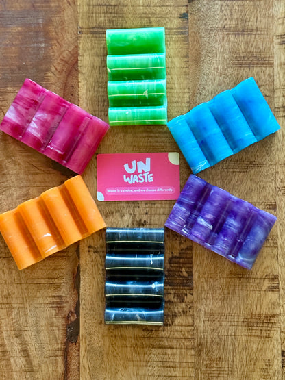 Six vibrant soap bars—pink, green, blue, purple, black, and orange—are arranged in a circle around a card that reads “Unwaste” on a wooden surface, inspiring the ultimate zero-waste bathroom setup. These colorful soaps look ready to be placed on your favorite Upcycled Soap Tray by Unwaste.